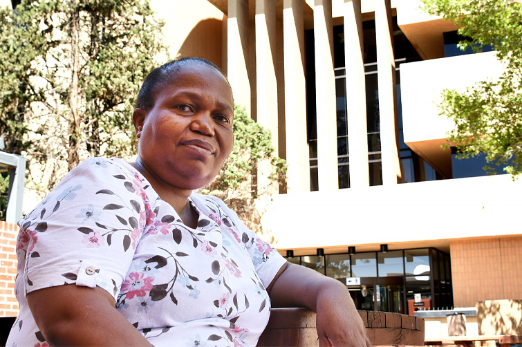 Nthabiseng Kotsokoane in front of the Sasol Library on the Bloemfontein Campus of the University of the Free State. She is wearing a pink blouse with floral patterns and leaning with her left elbow against a ledge whilst looking into the camera.